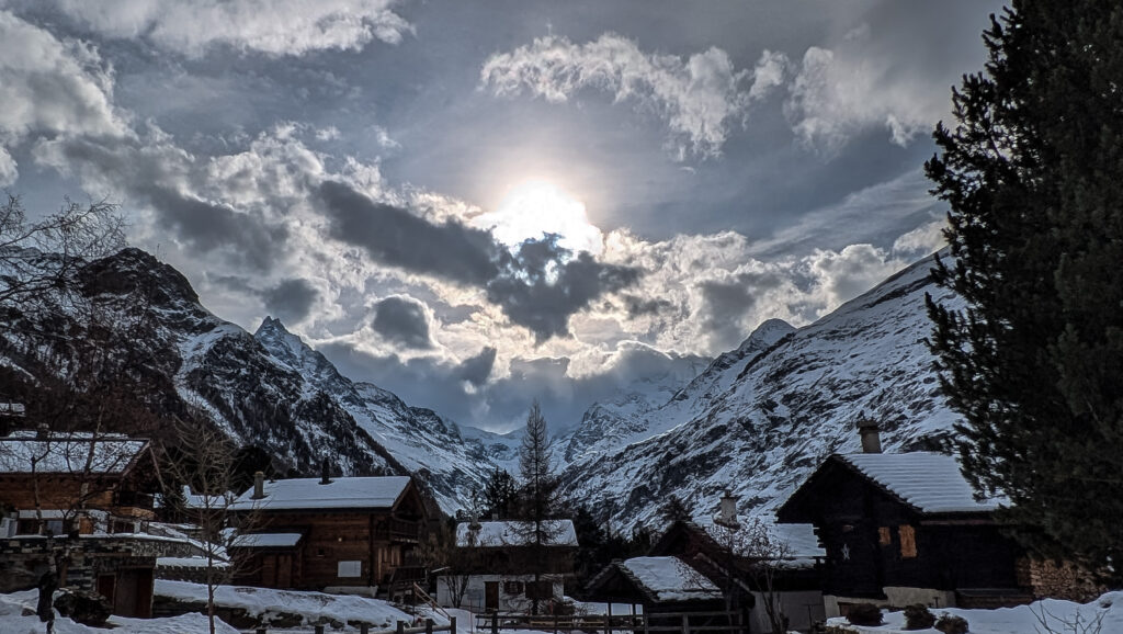 Vue des montagnes enneigées avec un ciel nuageux et le soleil entraînant des reflets sur les flocons de neige, encadrée par des chalets en bois.