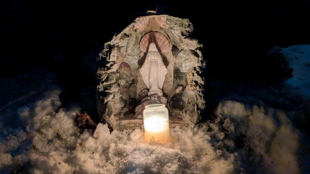 Statue de la Vierge Marie éclairée par une bougie, posée sur la neige et entourée de pierre.